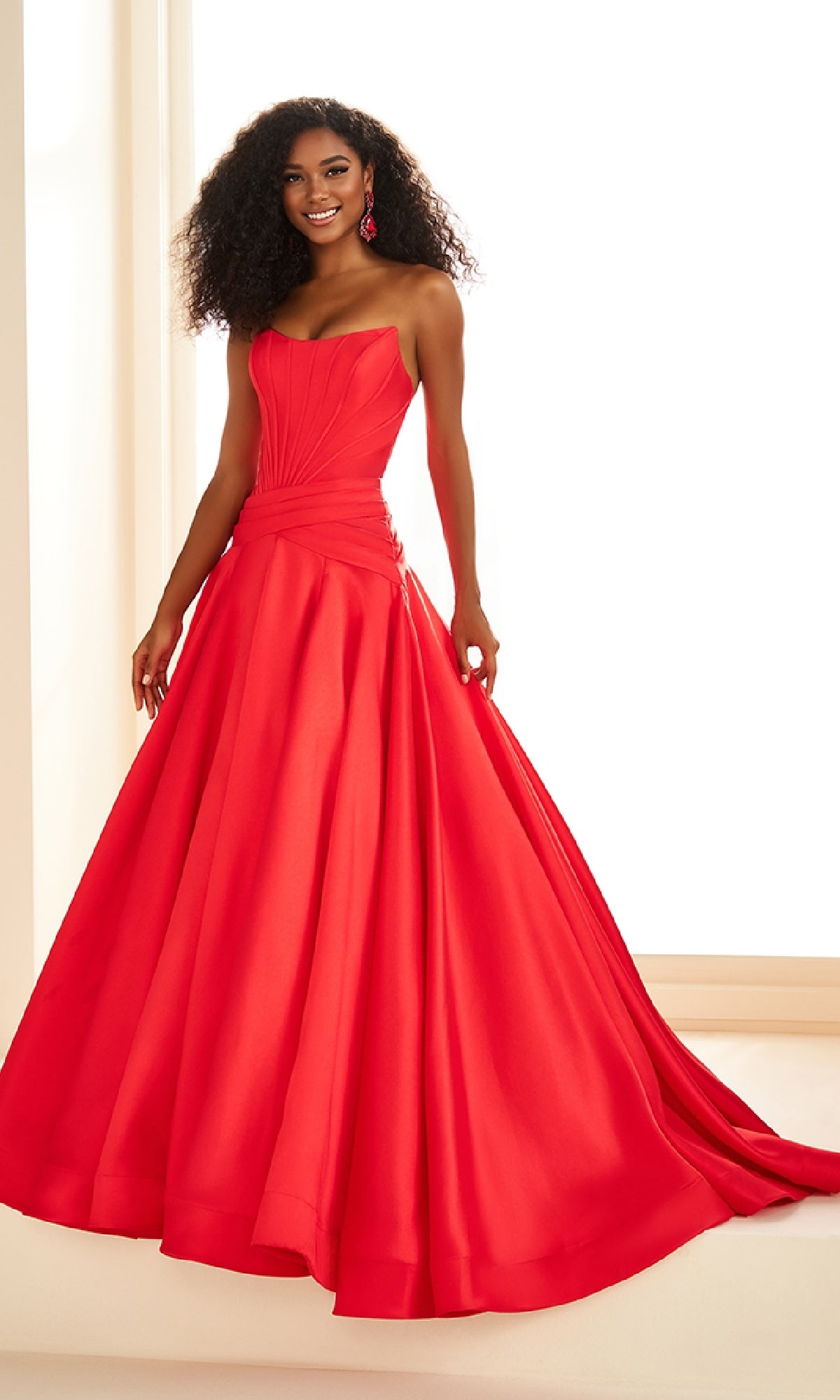 A woman wearing the Ellie Wilde EW36105 long prom dress—with a strapless mikado ball gown silhouette, corset bodice, and dropped waist—smiles at the camera indoors.